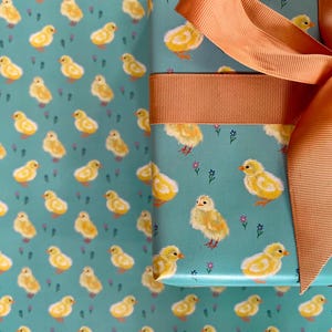 Peut inclure: Un cadeau emball&eacute; dans du papier turquoise orn&eacute; de motifs de poussins jaunes et de petites fleurs violettes. Le cadeau est nou&eacute; avec un ruban et un n&oelig;ud p&ecirc;che, id&eacute;al pour P&acirc;ques ou le printemps.
