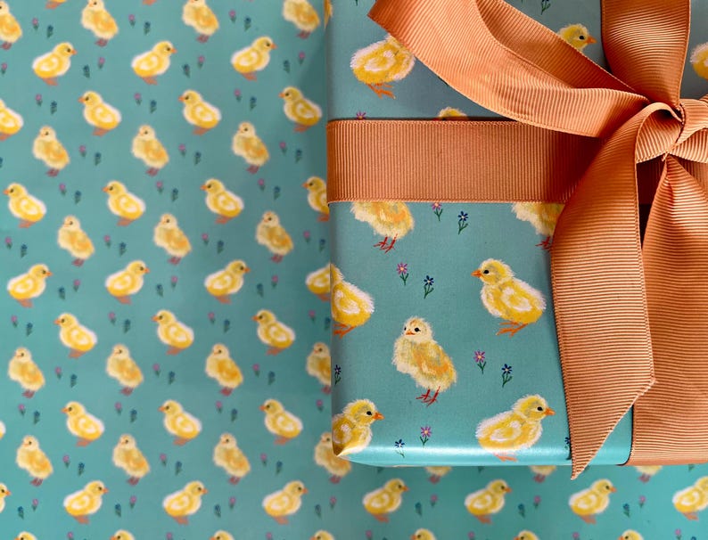 Peut inclure: Un cadeau emball&eacute; dans du papier turquoise orn&eacute; de motifs de poussins jaunes et de petites fleurs violettes. Le cadeau est nou&eacute; avec un ruban et un n&oelig;ud p&ecirc;che, id&eacute;al pour P&acirc;ques ou le printemps.