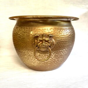 May include: A round brass planter featuring a hammered texture across its exterior surface. The vessel is adorned with a prominent, centered lion head relief ornament that holds a circular brass ring in its mouth, creating a decorative handle.