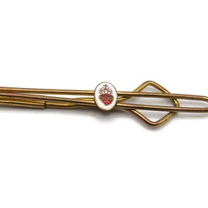 May include: Vintage gold-tone tie clip with an oval emblem. The emblem features a red heart with a crown of thorns, set against a white background. The tie clip has a classic design with a diamond-shaped accent.