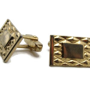 May include: A pair of gold-toned cufflinks with a geometric design. The cufflinks have a rectangular shape with a raised, textured border.