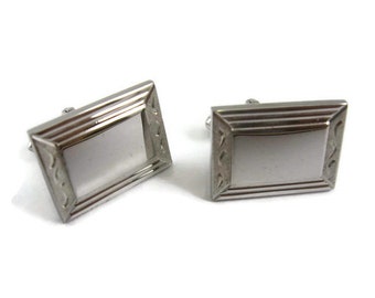 May include: A pair of silver-toned rectangular cufflinks. Each cufflink features a rectangular center with a textured border and decorative accents on the corners. The cufflinks are set against a white background.