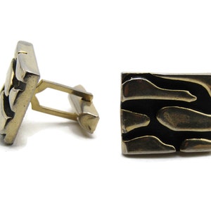 May include: A pair of square cufflinks with a black background and gold-colored abstract shapes. The cufflinks have a gold-colored metal frame and a gold-colored bar for securing them to a shirt cuff.