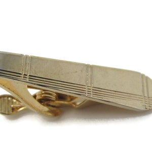 May include: A gold-toned tie clip with a textured, geometric design. The clip has a spring-loaded mechanism.