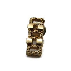 May include: A gold-colored, rectangular pin with a textured, brutalist design. The pin features three open rectangular sections with small, solid squares at the top and bottom of each section. The pin is set against a white background.