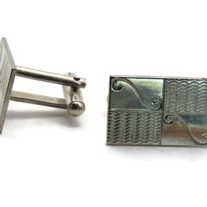 May include: Silver-toned cufflinks with a geometric design. One cufflink is rectangular with a four-part pattern of wavy lines and swirls. The other cufflink is square with a grid pattern. The cufflinks have a post and toggle closure.