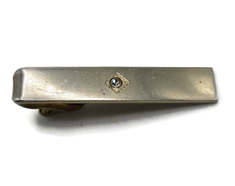 May include: Silver-toned tie bar with a small, clear gemstone set in a gold-colored, diamond-shaped frame. The tie clip has a rectangular shape with rounded edges and a clasp on the back.