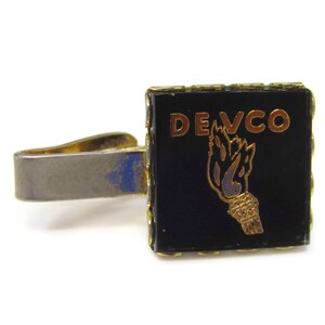 May include: Gold tie clip with a black background and the text "DEVCO" in gold letters. A gold torch design is below the text.