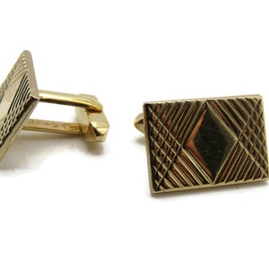 May include: A pair of gold-toned cufflinks. Each cufflink has a rectangular face with a textured, geometric design. The design features a diamond shape in the center, surrounded by a crisscross pattern. The cufflinks have a classic, elegant appearance.