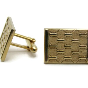May include: A pair of gold-toned cufflinks. One cufflink is angled, showing a rectangular face with a grid pattern. The other cufflink is face-up, displaying a rectangular face with a repeating geometric design. The cufflinks have a shiny finish.