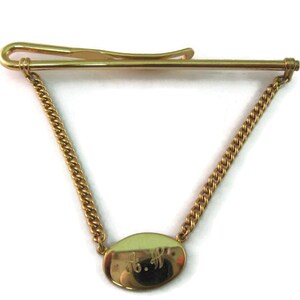 May include: Gold-toned tie clip with a chain and an oval pendant. The pendant has a light green enamel background and engraved initials. The clip is designed to secure a tie to a shirt.
