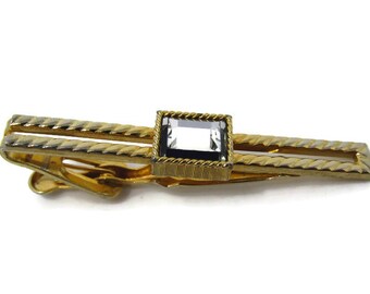 May include: A gold-toned tie clip with a rectangular, gray gemstone center. The clip has a textured, rope-like design along the edges and a hinged clasp. A classic accessory for formal wear.
