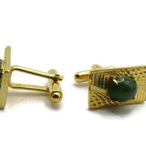 May include: A pair of gold-toned cufflinks with a square design and a textured surface. Each cufflink features a dark green, round stone. The cufflinks are set against a white background.