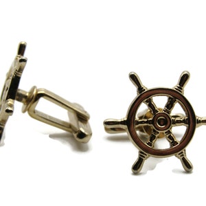 May include: Gold-toned cufflinks featuring a ship's wheel design. The wheel has a brown center and six spokes. The cufflinks have a classic, nautical theme and are set against a white background.