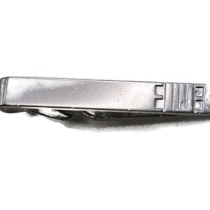 May include: A silver-toned tie clip with a rectangular design. The clip has a simple, classic style with a textured surface and a decorative end with the letters "EUE". The clip is designed to hold a tie in place.