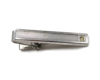 May include: Silver-toned tie clip with a rectangular design and a small, square, yellow gemstone. The clip has a textured, ridged pattern and a hinged clasp. A classic accessory.