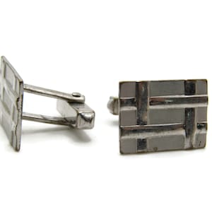 May include: A pair of silver-toned cufflinks with a rectangular shape and a woven design. The cufflinks are set against a white background, highlighting their metallic sheen and intricate pattern. These accessories are suitable for formal wear.