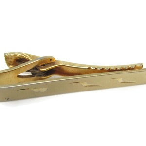 May include: Gold-toned tie clip with a textured, patterned design. The clip has a serrated edge and a spring-loaded mechanism.