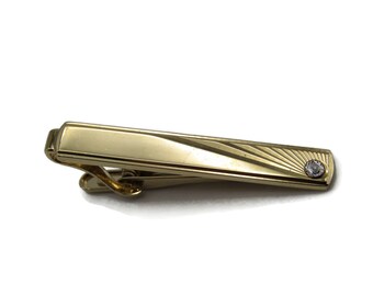 May include: A gold-toned tie clip with a rectangular design. The clip features a small, round, clear gemstone on one end and a series of engraved lines radiating from the gemstone. The clip is designed to hold a tie in place.