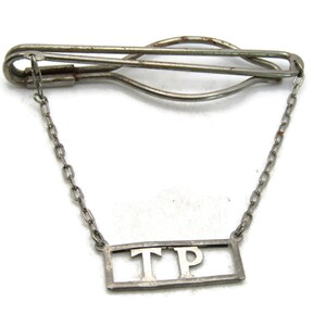 May include: A vintage silver-toned tie clip with a chain and a rectangular pendant. The pendant features the initials "TP" in a block letter style. The clip is designed to secure a tie to a shirt.