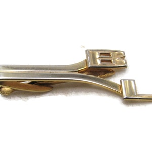May include: A gold and silver-toned tie clip with the letters "BL" in a stylized font. The clip has a rectangular shape with a curved design and a clasp on the back. The letters are in a block style.