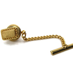May include: A gold-toned tie tack with a rectangular face and a decorative border. The tack is connected to a chain and a bar-shaped clasp. The design is simple and elegant, suitable for formal wear.