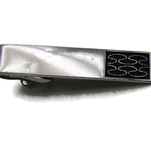 May include: Silver-toned tie clip with a rectangular black enamel inlay. The inlay features a repeating pattern of black, interconnected shapes outlined with white dots. A classic accessory for formal wear.