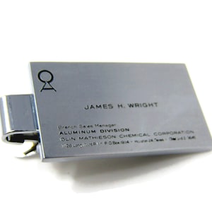 May include: A silver metal business card holder with a black logo and text. The text reads "JAMES H. WRIGHT, Branch Sales Manager, ALUMINUM DIVISION, OLIN MATHIESON CHEMICAL CORPORATION, 1/28 Long Island, P.O. Box 914, Hicksville, New York 11801".