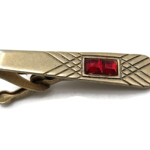 May include: Vintage gold-tone tie clip with a rectangular red jewel centerpiece. The clip features a geometric design and a clasp for securing to a tie. A classic accessory for formal wear.