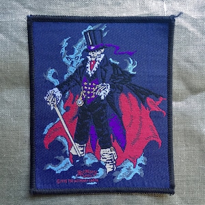 Puede incluir: Parche bordado con una figura oscura con un sombrero de copa y una capa, con un bastón blanco y un diseño rojo y morado. El texto "Alchemy Gothic" y "©1996 The Alchemy Carta" son visibles en la parte inferior.