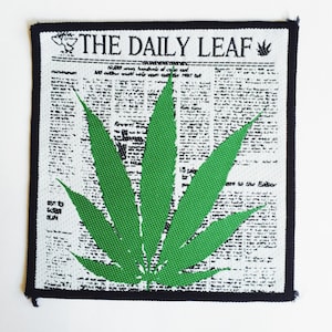 Puede incluir: Un parche blanco con ribete negro presenta una hoja de marihuana verde impresa sobre una impresión de periódico en blanco y negro. El texto "THE DAILY LEAF" está impreso en la parte superior del parche.