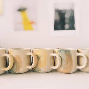 May include: A row of handmade ceramic mugs with rounded handles. The mugs have a beige base color with abstract watercolor-style designs in shades of yellow, green, and brown. The mugs are lined up on a white surface.