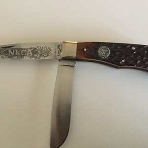 May include: A pocket knife with a partially open blade. The blade is engraved with "NKCA 1988" and decorative flourishes. The handle is brown with a textured pattern and a metal emblem. The knife is made by Camillus.