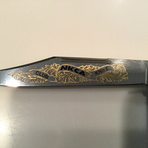 May include: A silver pocket knife with a gold engraved design featuring the text "NKCA Knife Club 1990".
