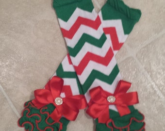 Op de afbeelding: Een paar beenwarmers met een groen en rood chevron-patroon met rode strikken en witte parelversieringen. De beenwarmers hebben een groene ruche-bies.