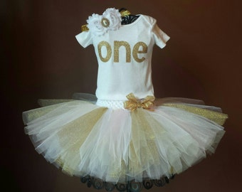 May include: A white tutu with gold and pink tulle layers, a white shirt with gold glitter lettering that says "one", and a white flower headband with gold accents.