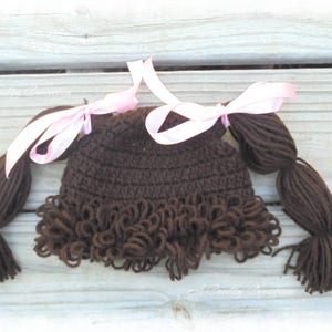 May include: A dark brown crocheted hat featuring yarn hair styled into two pigtails, each tied with a light pink satin ribbon. The hat includes a fringe of curly brown yarn along the bottom edge, mimicking a hairstyle with bangs and side pigtails.