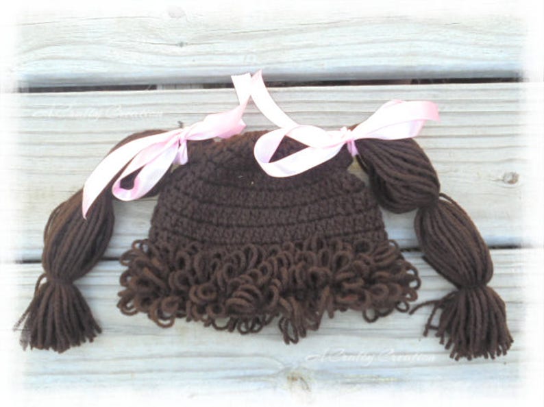 May include: A dark brown crocheted hat featuring yarn hair styled into two pigtails, each tied with a light pink satin ribbon. The hat includes a fringe of curly brown yarn along the bottom edge, mimicking a hairstyle with bangs and side pigtails.
