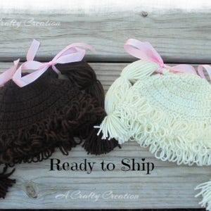 May include: Two crocheted hats with yarn hair, one in dark brown and one in cream. Each hat features looped yarn bangs and two side ponytails tied with light pink satin ribbons. The hats are designed to resemble hair with pigtails.