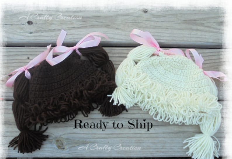 May include: Two crocheted hats with yarn hair, one in dark brown and one in cream. Each hat features looped yarn bangs and two side ponytails tied with light pink satin ribbons. The hats are designed to resemble hair with pigtails.
