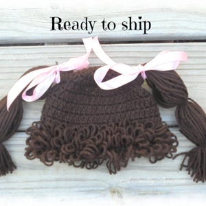 May include: A dark brown crocheted hat featuring yarn pigtails on each side, accented with light pink satin ribbons tied in bows. The hat has a textured, curly fringe along the bottom edge and the text Ready to ship is visible above the hat.