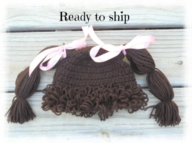 May include: A dark brown crocheted hat featuring yarn pigtails on each side, accented with light pink satin ribbons tied in bows. The hat has a textured, curly fringe along the bottom edge and the text Ready to ship is visible above the hat.