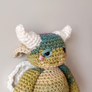 May include: A handmade crochet doll with a whimsical design. The doll features a multicolored body with shades of green, blue, and beige. It has white horns, blue eyes, and small pink cheeks. White crochet wings are attached to the back.