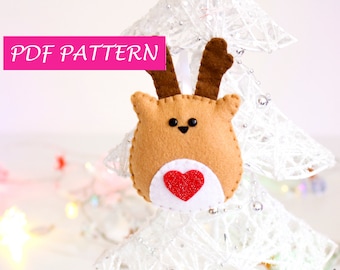 May include: A felt reindeer ornament with a red heart on its chest. The ornament is hanging from a white, sparkly Christmas tree. The text "PDF PATTERN" is displayed in pink at the top of the image.