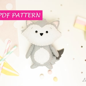 May include: PDF pattern for a grey felt wolf toy with black eyes and a white belly. The pattern is available for download. The image also includes a pink and yellow striped candy, a white marshmallow, and gold glitter stars.
