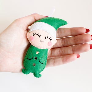 May include: A green felt elf ornament with a white felt hat and a smiling face. The elf has gold glitter stars on its green felt outfit.