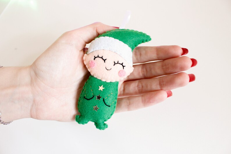 May include: A green felt elf ornament with a white felt hat and a smiling face. The elf has gold glitter stars on its green felt outfit.