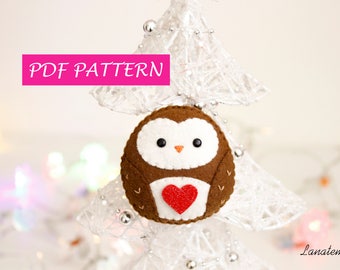 May include: PDF pattern for a felt owl ornament with a red heart. The owl is brown with white facial features and is hanging on a white, sparkly Christmas tree.