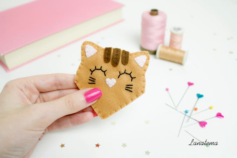 May include: A felt cat bookmark with brown stripes, pink ears, and black eyes. The bookmark is being held by a hand.