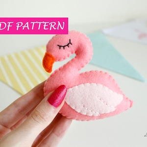 Può includere: Un fenicottero rosa in feltro con la pancia bianca in feltro e occhi neri. Il fenicottero è tenuto in una mano. Il testo "PDF PATTERN" è in lettere rosa su sfondo bianco.